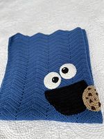 Full folded view of the blue crocheted blanket with chevron pattern, showing Cookie Monster face with eyes, mouth, and a cookie appliqued on one corner.