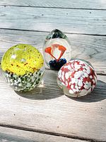 Three Mid-Century Modern glass paperweights shown on wooden surface: yellow circular, red and white rounded, and orange mushroom-shaped egg piece.