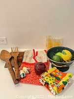 Photo shows large wooden spoon and fork with natural finish, 2 teak egg cups, colorful printed cotton towels, salt and pepper pinches, ceramic fruit piece, ceramic apple, and 3 glasses behind a black colander holding 2 lemons and an avocado.