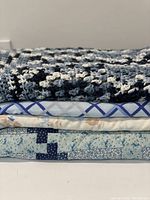Stacked view of handmade quilt, hand crochet throw, and two flat sheets showing blue-toned patterns and textures.