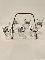 Six vintage French hand-painted shot glasses arranged around and inside gold-tone metal carrying rack with red grips on handle and legs.