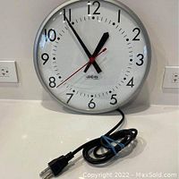 Front view of the vintage wall clock showing black hour and minute hands, red second hand, white face with bold black numbers, and attached power cord.