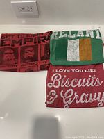 Photo of three folded vintage T-shirts: Star Wars Empire character chart on red shirt, Ireland flag shirt, and red biscuits and gravy text shirt.