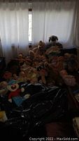 Dark photo showing a collection of plush dolls and toys arranged on a bed with a curtain backdrop. Visible stuffed animals include various bears and character toys, positioned sitting and lying down.