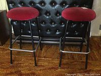 Pair of barstools with black metal frames and red padded seats shown from front view on brown carpet in front of black tufted surface.