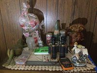Overview showing all items on lace cloth on wooden surface against wood panel wall. Includes binoculars, ET figurine, bottle, cans, toy boat, small house, calculator, frosted figurine, Kermit frog statue, and paper clips boxes.