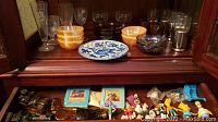 Wide shot showing carnival glass bowls, stemware, metal-sleeved glasses, Simpsons toys, miniature Christmas figurines, and Avon bottles spread across two shelves