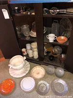 White glass serving bowl with matching ruffled platter and ladle in front of cabinet with assorted coffee mugs, platters, candy bowls, metal grater and hand mixer inside.