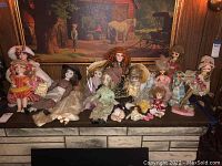 Photo showing sixteen dolls of porcelain and plastic faces posed on a black bench beneath a framed painting, variously dressed in vintage and classic clothing, some seated, some standing.