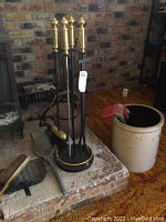Photo showing brass-topped fireplace tool set on black stand next to ceramic jug with gloves inside and black metal umbrella stand
