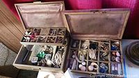 Two vintage jewelry boxes open showing compartments filled with assorted costume jewelry such as earrings, rings, necklaces, brooches, and pins, along with a wristwatch and sewing supplies.