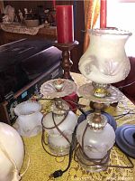 Photo showing two frosted white glass table lamps with one lamp missing its shade and wiring exposed, a wooden candle holder with a red candle, and parts of other lamps and decor on a table.