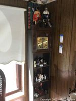 Wide view of a shelving unit holding two nutcrackers on top, one in original box, multiple smaller figurines in the shelves below, against wooden paneled wall near window.