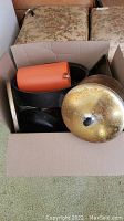 Box containing various pots, pans, and lids including an orange Tupperware pitcher and metal cookware.