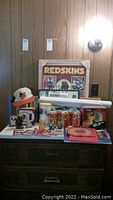 View of Baltimore Orioles baseball cap, Redskins framed poster, collectible cards, NFL goalpost mug, owl and frog figurines, book and Dean Martin 45, all arranged on wooden dresser with wood-paneled wall background.