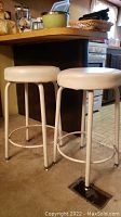 Two white vinyl top counter stools with metal legs placed on carpeted floor by a kitchen counter.