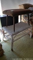 Wicker table with lower shelf, white painted, showing wear and some dirt at base. Ottoman stool on top with wooden legs and brown fabric upholstery.