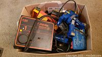 Box containing various electric train transformers, HO scale train cars, engines and spare pieces with visible power cords and transformers mainly blue and orange in color.