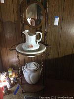 Full view of the washstand showing wooden frame with oval mirror and the ceramic pitcher on top, lidded jar below, and basin on stand.
