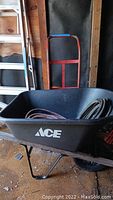 Photo showing the Ace wheelbarrow with two coiled hoses inside and the hand truck behind it.