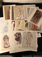 Overhead view of about 15 prints spread on black surface showing varied historical and fantasy costume illustrations.