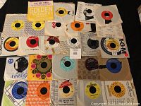 Top down view of 25 assorted 45 RPM vinyl records displayed on fabric, showing variety of record labels and artist names.