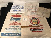 Five cloth sacks laid out side by side, four with distinct advertising prints and one blank sack, displayed on a black surface.