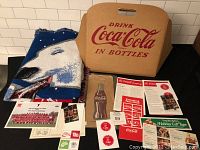 Photo showing the folded Coca-Cola polar bear blanket, the folded NOS cardboard Coca-Cola stadium seat, and various paper advertising items arranged in front.