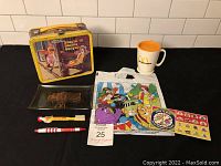 Photo showing entire lot items on black background including lunch box, coffee cup, glass tray, litter bag, game spinner, toothbrush, and pizza pen.