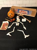 Full view of skeleton puppet with detached limbs laid out, two noisemakers, two shell out bags, original box, and pumpkin light bulb on black cloth background.