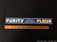 Front view of rectangular porcelain Purity Flour advertising sign next to a ruler for scale showing dimensions about 13 inches in length and 1.5 inches in height