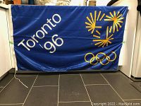 The full front view of the blue flag showing the markings 'Toronto 96', yellow stylized sunburst logo and Olympic rings in yellow, displayed against a black tiled floor.