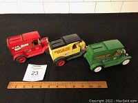 The three metal advertising truck banks aligned on a black surface next to a ruler showing approximate size, labels clearly visible: Red Rose Tea in red truck, Home Hardware in yellow truck, Canada Dry in green truck.