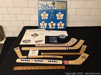 Toronto Maple Leafs collectible lot laid on black surface showing 5 mini souvenir hockey sticks with signatures, business envelope, game program, Tim Hortons puck, and 1964/65 Let’s Talk Hockey vinyl record album cover.