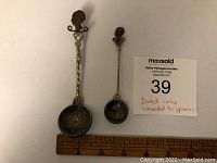 Photo showing two souvenir spoons placed next to a ruler for size reference, both made from Dutch silver coins with decorative tops.