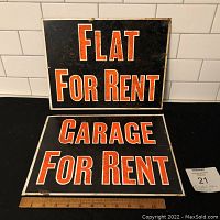 Two vintage rectangular metal rental signs, one 'Flat For Rent' and one 'Garage For Rent', placed on a black surface with ruler for scale, against white subway tile background.
