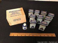Photo of the full original box containing nine small blue and white Fasteeth denture powder trial sample tins, one tin removed and placed beside the box with a ruler for scale.