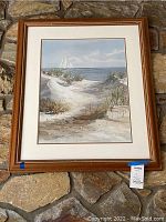 Framed painting showing beach dunes, grass, and a sailboat with wooden frame and white matting.