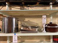 Large stainless steel stock pot, multiple bakeware pans including roasting pan and baking trays, white immersion blender and brown Dutch oven with lid on basement shelf