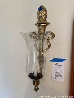 Side view of one brass-like decorative wall sconce showing clear etched glass hurricane globe, white taper candle, pineapple finial at top, and curled ribbon detail wrapping around the base.