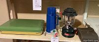 Lot displayed on basement shelf showing camping stove, blue thermos, Coleman lantern, and binoculars in box