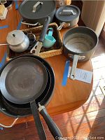 Overview of all cookware items on a wooden table including pans, pots, a glass baking dish in a basket, and a cocoa pot with blue kitchen measuring cups visible.