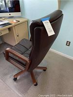 Side angle of brown leather desk chair showing wheeled base and wooden armrest, clear desk mat below chair.