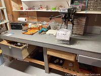 Wide shot showing the Central Machinery drill press mounted on workbench with drawers open and assorted tools and containers on work surface.