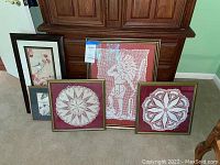 Overview of all five framed artworks arranged on carpet in front of wooden furniture showing variation in crocheted and printed art, different frame styles and sizes.