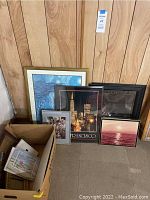 Photo showing assorted framed art leaning against wood paneled wall, a box of prints on floor.