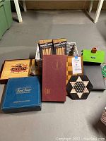 Overview photo of assortment of boxed and unboxed games including Mexican Train Dominoes, Trivial Pursuit, Chinese Checkers board, checkerboard with chess pieces and wooden boxes and trays.