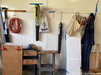 Full view of garage wall with Ryobi leaf blower and battery charger, coveralls on hanger, winter windshield cover hung on foam boards, rope and other items.