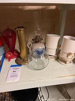 Shelf with brass lamp base, country style hurricane lamp, glass globe, white portable lamps, and red hummingbird feeder.