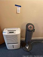 Frontal view of the white Frigidaire humidifier and adjacent Lasko space heater standing on the floor against a beige wall.